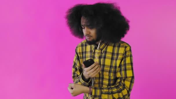 Uomo afroamericano positivo che usa il telefono per ascoltare musica nelle cuffie su sfondo viola. Concetto di emozioni — Video Stock