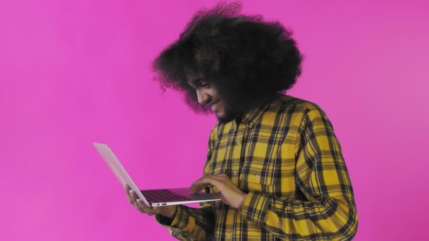 Hombre afroamericano escribiendo en el portátil y sonriendo sobre fondo púrpura. Concepto de emociones — Vídeos de Stock