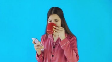 Pijamalı mutlu kız çay ya da kahve içiyor ve stüdyoda mavi arka planda telefon kullanıyor.