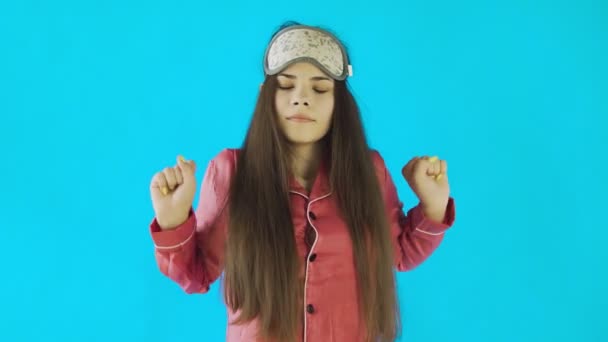 Jong mooi meisje in pyjama geeuwen en stretching op blauw achtergrond in studio — Stockvideo