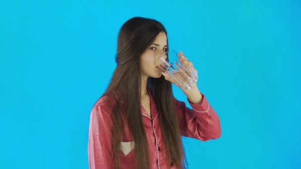 O adolescentă caucaziană bea un pahar cu apă. O tânără bea apă din sticlă pe fundal albastru în studio — Videoclip de stoc