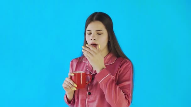 Jeune femme en pyjama boit du thé ou du café sur fond bleu en studio — Video