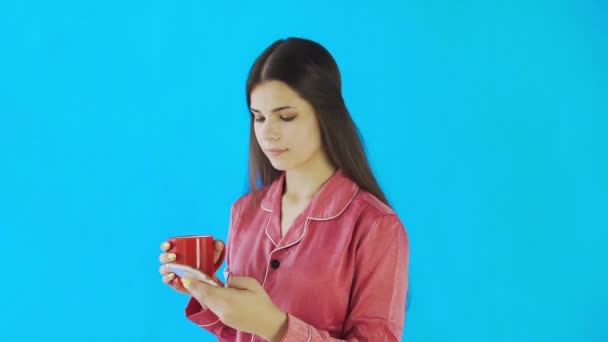 Gelukkig meisje in pyjama is het drinken van thee of koffie en het gebruik van telefoon op blauwe achtergrond in de studio — Stockvideo