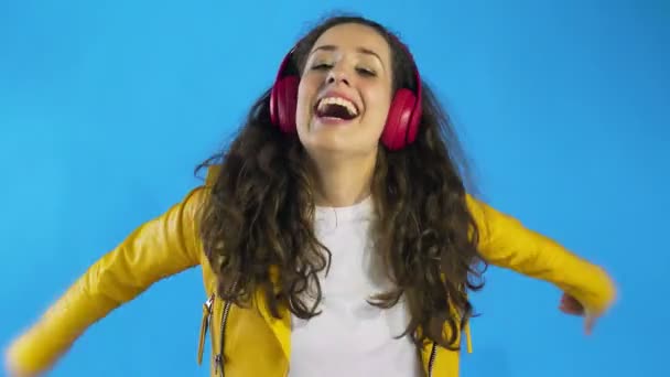 Молода жінка в жовтій куртці танцює під час прослуховування музики на навушниках у студії з синім тлом . — стокове відео