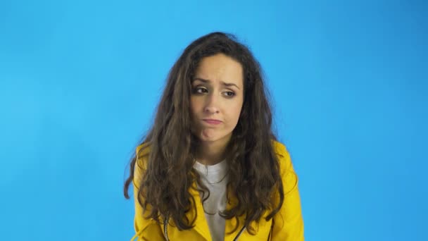 Triste mujer joven hermosa reflexiva en chaqueta amarilla de pie en el estudio con fondo azul . — Vídeos de Stock