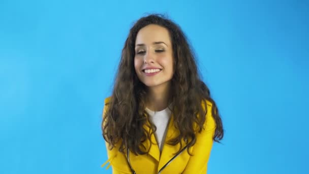 若いです女性で黄色のジャケットありますかむ唇とウインク目でスタジオで青の背景. — ストック動画