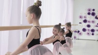 Çalışkan genç bale dansçıları plie ve battement tendu yaparken bayan öğretmenleri yanlış pozisyonları düzeltiyor ve talimatlar veriyor..