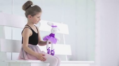 Elinde oyuncak olan küçük kız bale sınıfında bankta oturuyor.