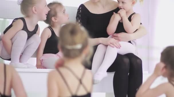 Giovane insegnante di balletto seduto sulla panchina nella scuola di balletto con bambine in body neri e comunicare con loro — Video Stock