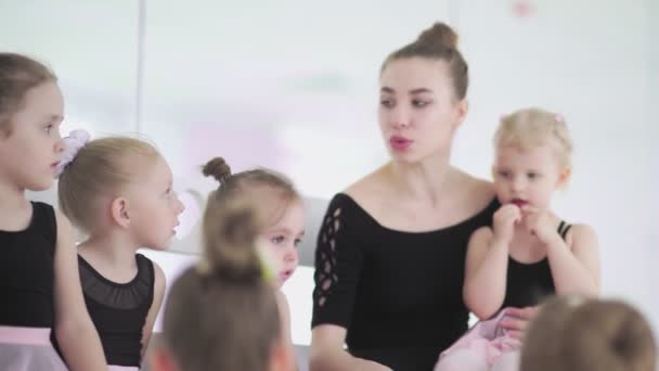 Junge Ballettlehrerin sitzt mit kleinen Mädchen in schwarzen Trikots auf Bank in Ballettschule und kommuniziert mit ihnen — Stockvideo