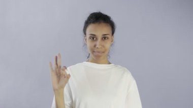 Şirin Afro-Amerikalı Kız iki başparmağıyla iyi görünüyor ve stüdyodaki gri arkaplanda gülümsüyor.