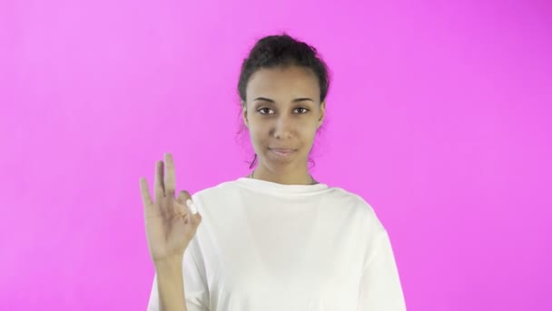 CuteAfro-American Girl показує себе в порядку з двома пальцями і посміхається на рожевому фоні в студії — стокове відео