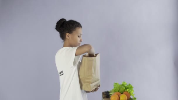 Gelukkig vrouw laden papieren zak met biologische gezonde groenten en fruit op grijze achtergrond — Stockvideo