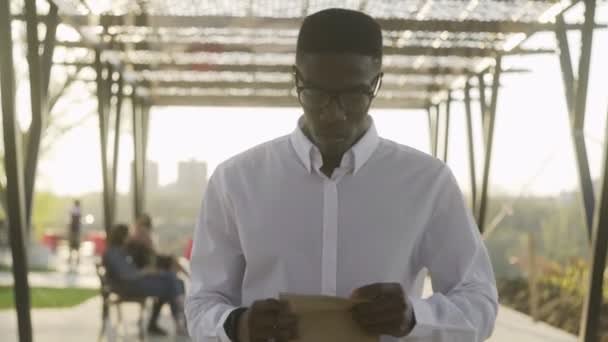 Афроамериканский бизнесмен в белой рубашке пьет кофе в парке возле офиса — стоковое видео
