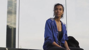 Otobüs durağında otobüs bekleyen ve eğitimden sonra kulaklıkla akıllı telefondan müzik dinleyen genç bir kadın.