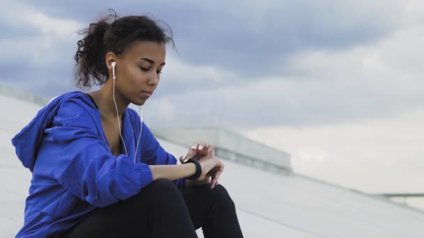 Mulher afro-americana esportiva em sportswear ouvir música e monitorar seu progresso em um smartwatch — Vídeo de Stock