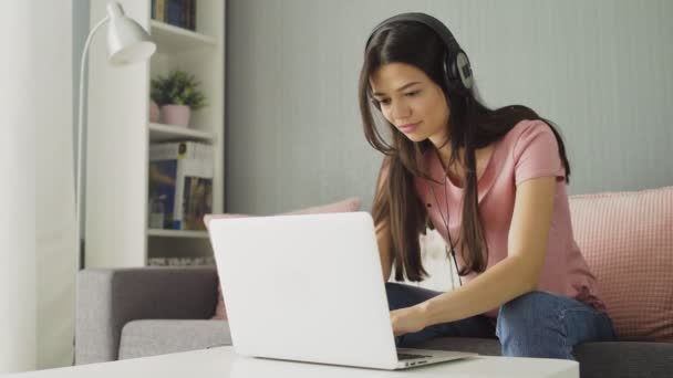Mosolygó fiatal nő fülhallgatóval zenét hallgatni és szörfözni az interneten otthon — Stock videók