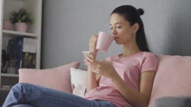 Akıllı telefonu dokunmatik ekranla kullanan ve sabahları kahve içen genç bir kadın.