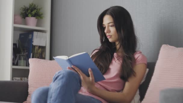 집에서 소파에 앉아 책을 읽고 있는 아름다운 젊은 여성 — 비디오