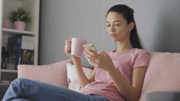 自宅でソファでリラックスしているスマートフォンを使って美しい若い女性オンラインテキストメッセージ飲料コーヒー快適なライフスタイルを楽しむ — ストック動画