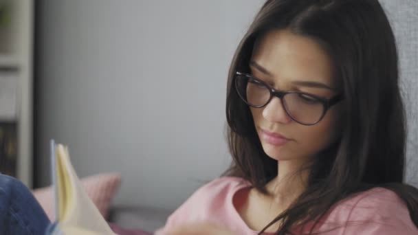 Mooie vrolijke studente met bril die een boek leest op de bank van een modern huis. Kijken. Terug naar school. Onderwijsuniversiteit. — Stockvideo