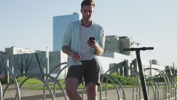 晴れた日に公園に黒いスタイリッシュな電動スクーターが立っているハンサムな男。スマートフォンを使う男 — ストック動画