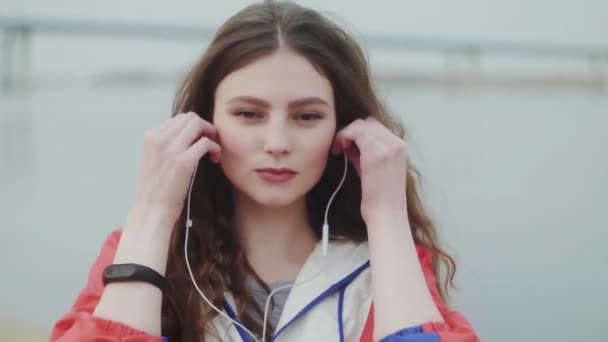 Jonge vrouw lokking op de camera en het dragen van een koptelefoon voor muziek motivatie voor cardiotraining, rivier op de achtergrond — Stockvideo