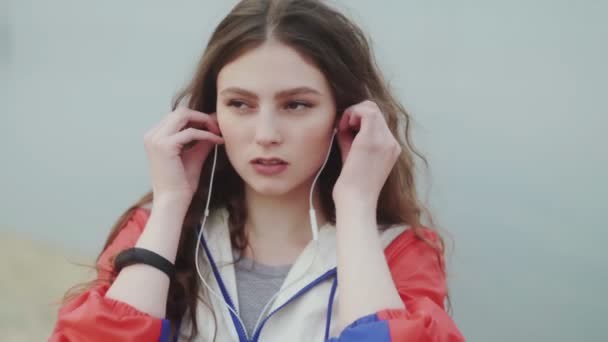 Mujer joven está usando auriculares para la motivación musical para el entrenamiento cardiovascular, río o mar en el fondo — Vídeos de Stock