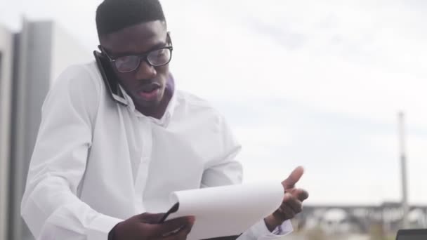 Занятой афроамериканский бизнесмен в очках разговаривает по мобильному телефону и проверяет документы в городском парке — стоковое видео