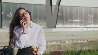 Güzel bir iş kadını cep telefonuyla konuşurken kruvasan yiyor ve dışarıda gülümsüyor.