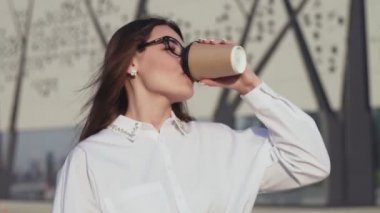 Gülümseyen zarif kadın elinde kahveyle şehir parkında yürüyor.