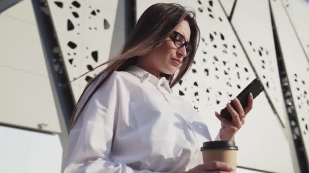 Schöne Geschäftsfrau mit Brille, die aufs Smartphone schaut und Kaffee trinkt. Modernes Bauen im Hintergrund — Stockvideo