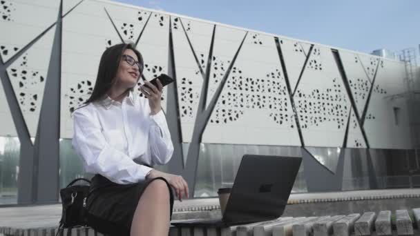 Mulher de negócios está gravando mensagem de voz enquanto está sentado no parque com seu caderno — Vídeo de Stock