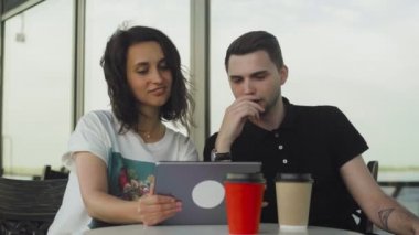 Bir kafede genç bir adam ve bir kadın. Tablete bakıp konuşuyorlar.