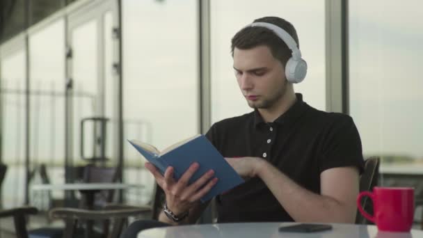Beau homme écoutant de la musique sur écouteurs et livre de lecture — Video