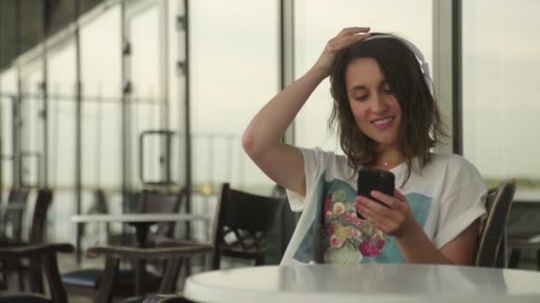 Молода жінка в навушниках розслабляється в кафе і слухає музику — стокове відео