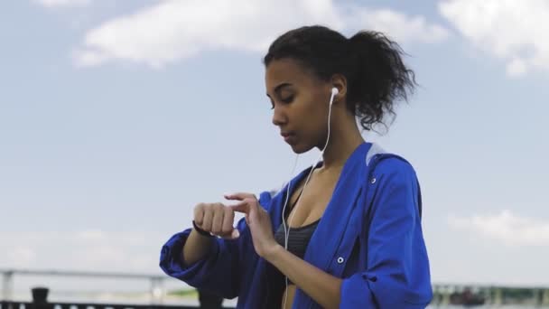 Runner ung afro-amerikansk kvinna kör och tränar utomhus med fitness tracker och kör app bärbar teknik med smartphone och hörlurar. Kvinnor som använder träningsarmband. — Stockvideo