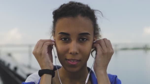 Jonge Afro-Amerikaanse vrouw kijkt naar de camera en het dragen van een koptelefoon voor muziek motivatie voor cardiotraining, rivier op achtergrond — Stockvideo