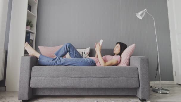Ελκυστική νεαρή γυναίκα πληκτρολογώντας στο έξυπνο τηλέφωνό της, ενώ βρίσκεται στον καναπέ στο σπίτι. Ψύξη γυναίκα χρησιμοποιώντας έξυπνο τηλέφωνο, κουβέντα, σερφάρισμα στο διαδίκτυο — Αρχείο Βίντεο