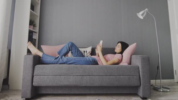 Красивая молодая женщина держит в руках смартфон и печатает на его экране, разговаривая в гостиной дома . — стоковое видео
