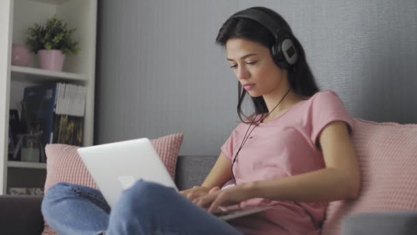 Young blonde woman with headphones sitting on sofa and working on laptop in the morning — Stock Video