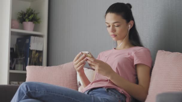 Mladá okouzlující brunetka žena sedí na šedé pohovce s růžovými polštáři, poslouchá hudbu na chytrém telefonu přes bílá sluchátka — Stock video