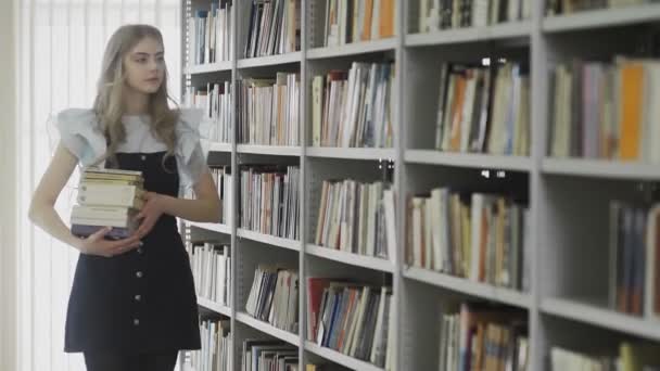 Hermosa joven estudiante rubia caminando en la biblioteca universitaria con muchos libros en las manos — Vídeo de stock