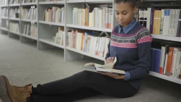 Ung afrikansk amerikansk kvinna läser bok sittandes på golvet i universitetsbiblioteket. — Stockvideo
