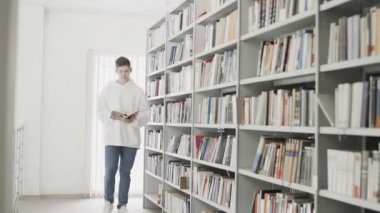 Kitap rafları arasında elinde kitapla dolaşan yakışıklı genç bir öğrenci.