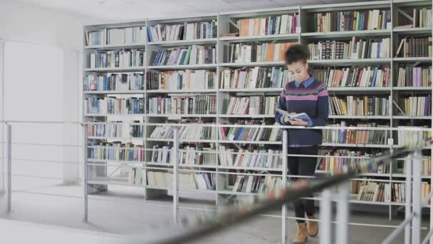 Jovem mulher americana africana bonita está lendo livro whlie de pé antes das prateleiras na biblioteca — Vídeo de Stock