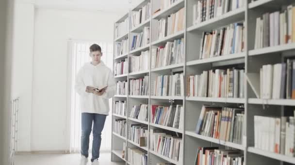 Guapo joven estudiante caminando entre estante de libros con libro en las manos — Vídeo de stock