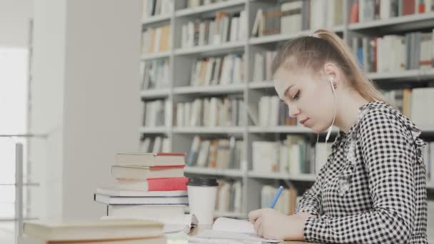 Konsentrert jentestudent med hodetelefoner som forbereder seg til eksamen og skriving mens hun sitter ved universitetsbiblioteket – stockvideo