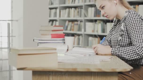 Dolly πλάνο του μαθητή κορίτσι με ακουστικά προετοιμασία για τις εξετάσεις και γράφοντας σημειώσεις, ενώ κάθεται στο τραπέζι στη βιβλιοθήκη του πανεπιστημίου — Αρχείο Βίντεο