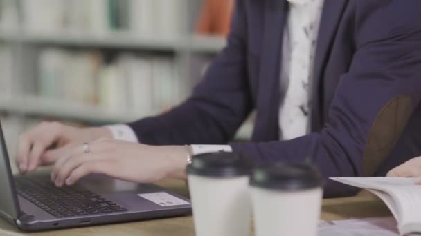 Joven hombre guapo se sienta y trabaja en el ordenador portátil en la biblioteca. Tazas de café en la mesa — Vídeo de stock